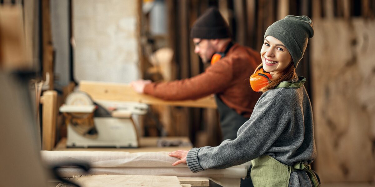 Couple of cheerful carpenters with wooden details in hands working together near workbench in professional workroom